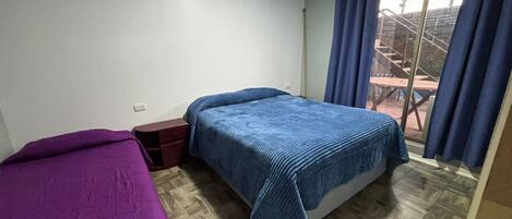 A bedroom with two beds, a wooden floor, a small bedside table, and a window with curtains.