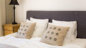 Un lit bien fait, avec une literie blanche et des coussins beiges ornés de motifs circulaires, une lampe sombre posée sur une table de chevet en bois, et un tableau floral encadré accroché au mur.