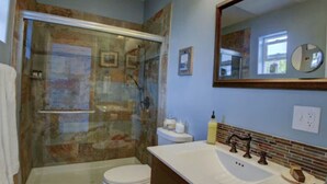 Un baño con ducha envuelta en vidrio, inodoro blanco, tocador con espejo y lavamanos.