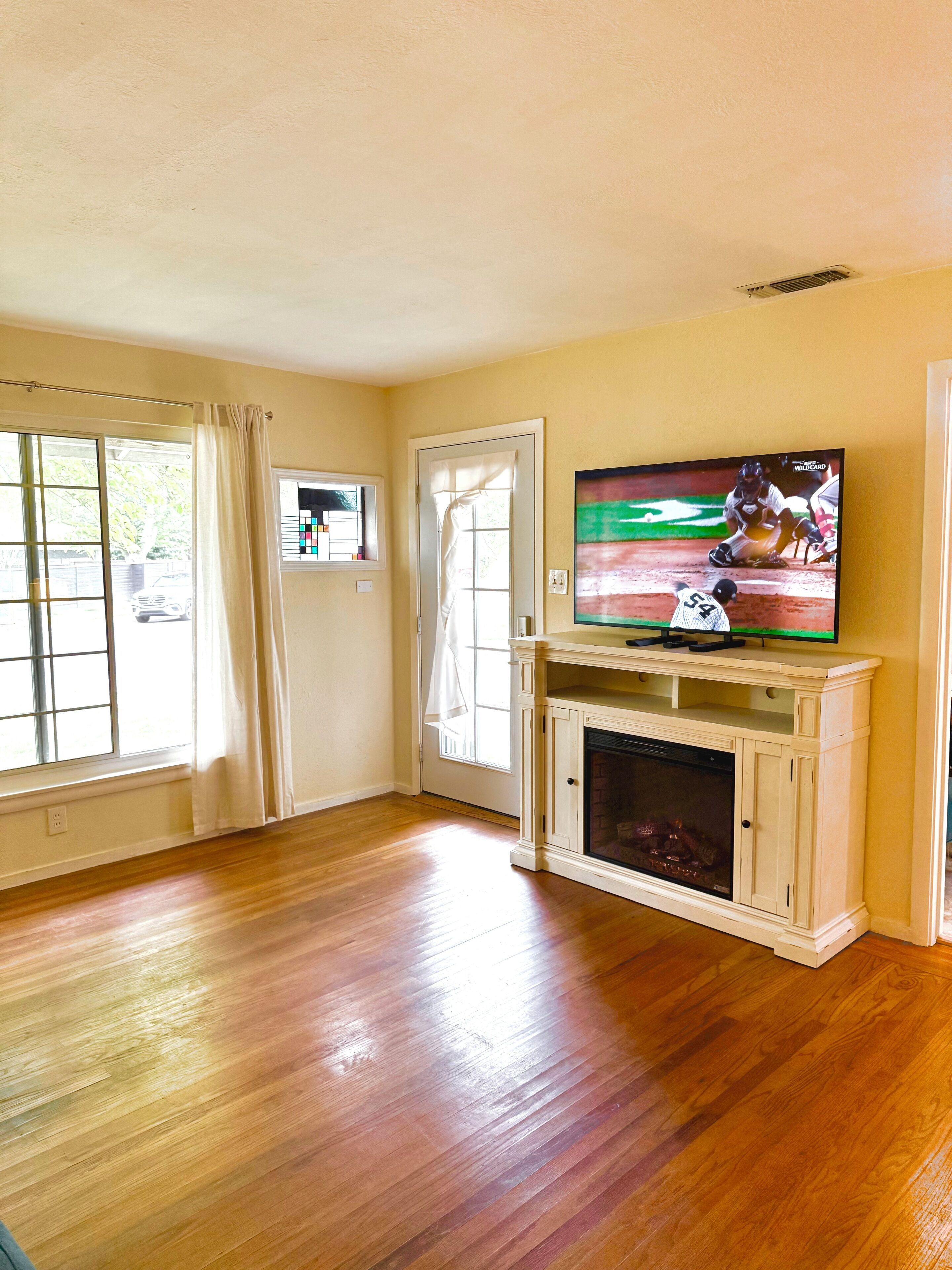 A living room with wooden flooring, a fireplace, and a television displaying a baseball game.