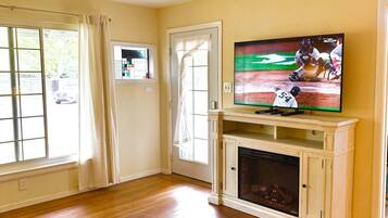 A living room with wooden flooring, a fireplace, and a television displaying a baseball game.