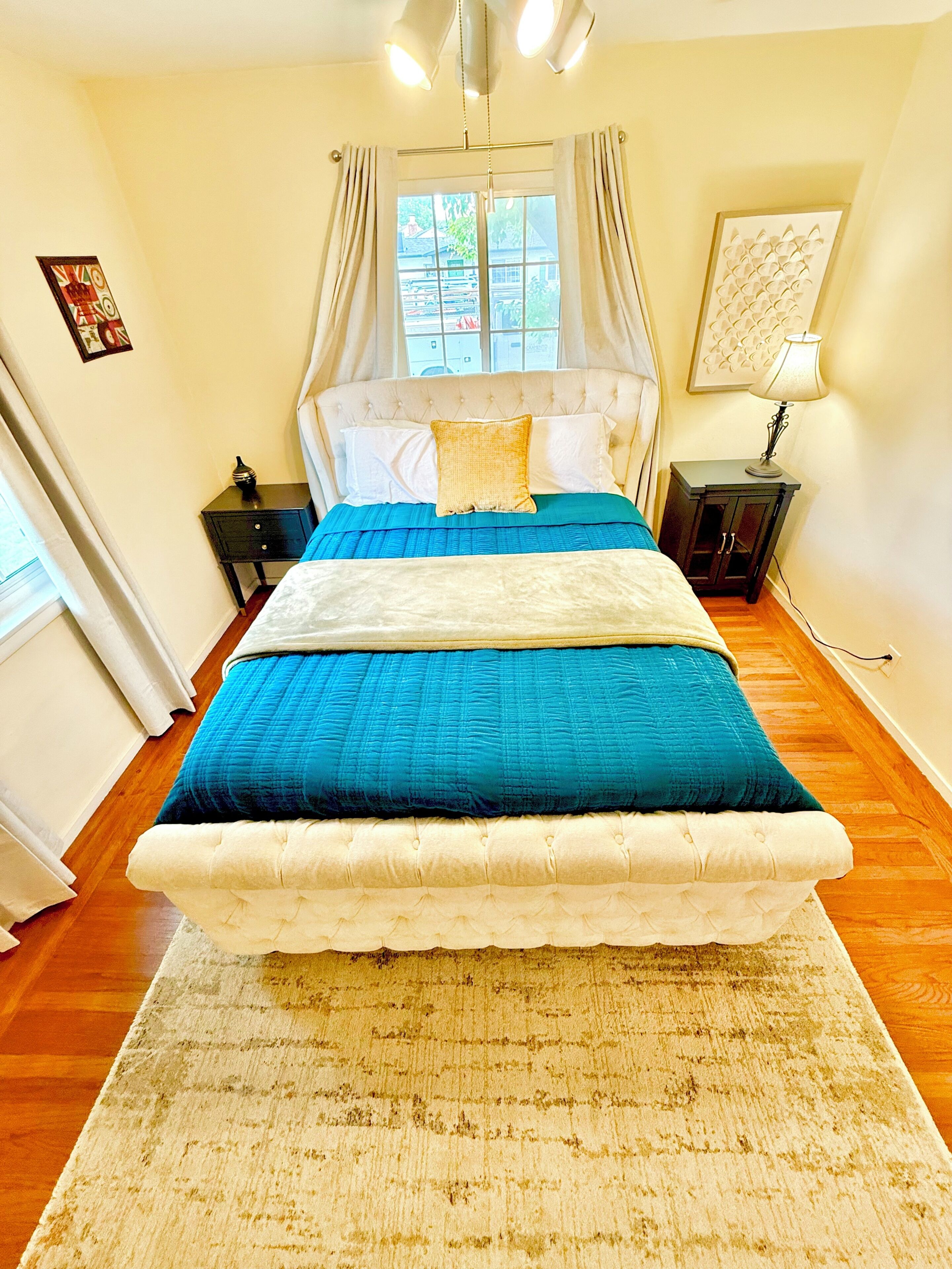 Une chambre à coucher comprenant un lit, une table de chevet, une lampe, une fenêtre avec des rideaux et une œuvre d’art accrochée au mur.