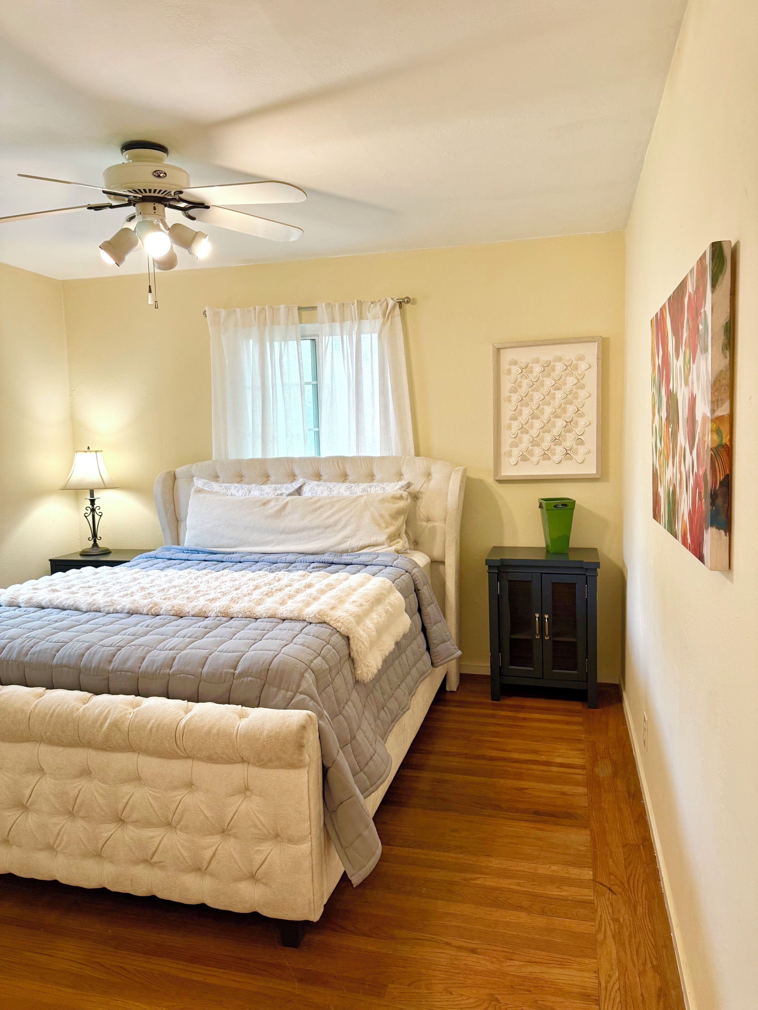 A bedroom with a bed, a ceiling fan, a nightstand, a lamp, a window with curtains, and a wall-mounted artwork.