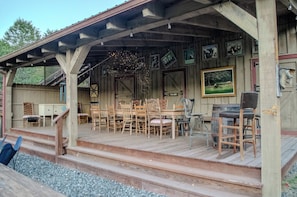 Um deck de madeira com área de estar coberta, cadeiras de madeira, uma mesa e um banquinho de bar.