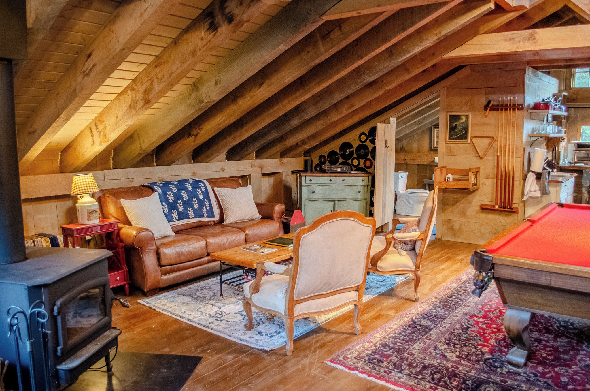 A cozy attic room with a wooden ceiling, a brown leather sofa, a pool table, a fireplace, and a dining area with a red lamp.