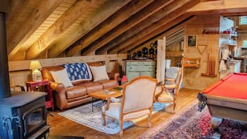 A cozy attic room with a wooden ceiling, a brown leather sofa, a pool table, a fireplace, and a dining area with a red lamp.