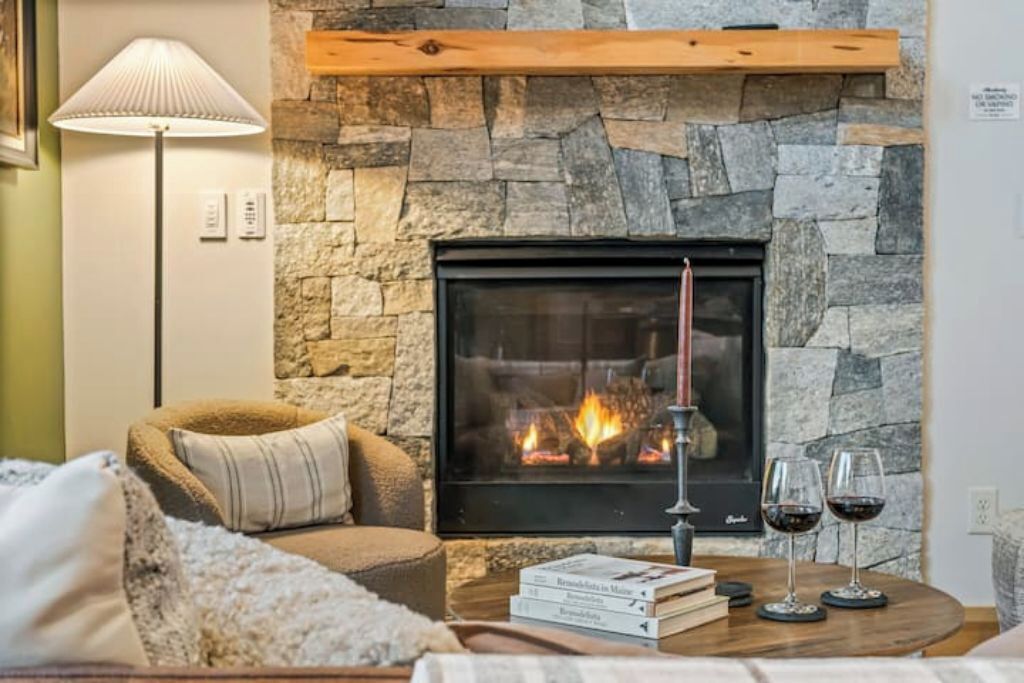 A cozy living room with a stone fireplace, a comfortable armchair, a floor lamp, and a small table with books and glasses of wine.