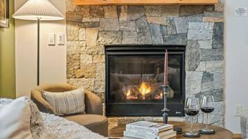 A cozy living room with a stone fireplace, a comfortable armchair, a floor lamp, and a small table with books and glasses of wine.