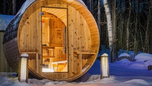 Avoinna olevalla ovella varustettu puinen mökki, jonka ympärillä on lunta ja jonka taustalla näkyy metsä.