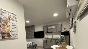 Een compacte keuken met moderne apparatuur, een eethoek met een gele stoel, en een bed met gestreepte beddengoed.