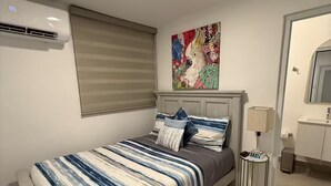 Un dormitorio con cama, ventana con persianas, aire acondicionado de pared, una obra de arte colorida, mesita de noche con lámpara y baño con espejo y lavamanos.