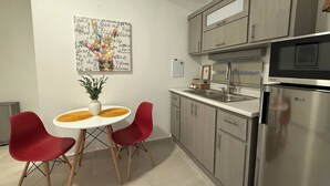 A modern kitchen with a stainless steel refrigerator, a microwave, and a sink. There is a dining table with two red chairs and a vase with a plant. A colorful welcome sign is on the wall.