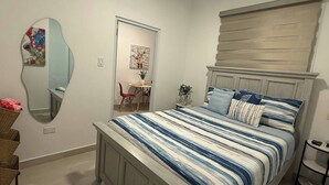 A bedroom with a bed, a mirror, a chair, a small table with a plant, and a shelf.