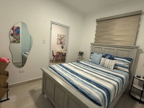 A bedroom with a bed, a mirror, a chair, a small table with a plant, and a shelf. - A&E L.A. Rental Apartments #4 (Carolina)
