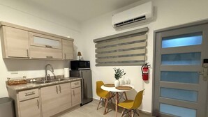 A compact kitchen with a microwave, sink, and cabinets. There is a small dining table with yellow chairs and a potted plant. A fire extinguisher is mounted on the wall. - A&E L.A. Rental Apartments #2 (Carolina)