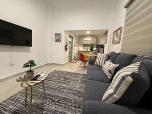 A modern living room with a grey sofa, a small side table with a plant, and a striped rug. A flat-screen TV is mounted on the wall. - A&E L.A. Rental Apartments #6 (Carolina)