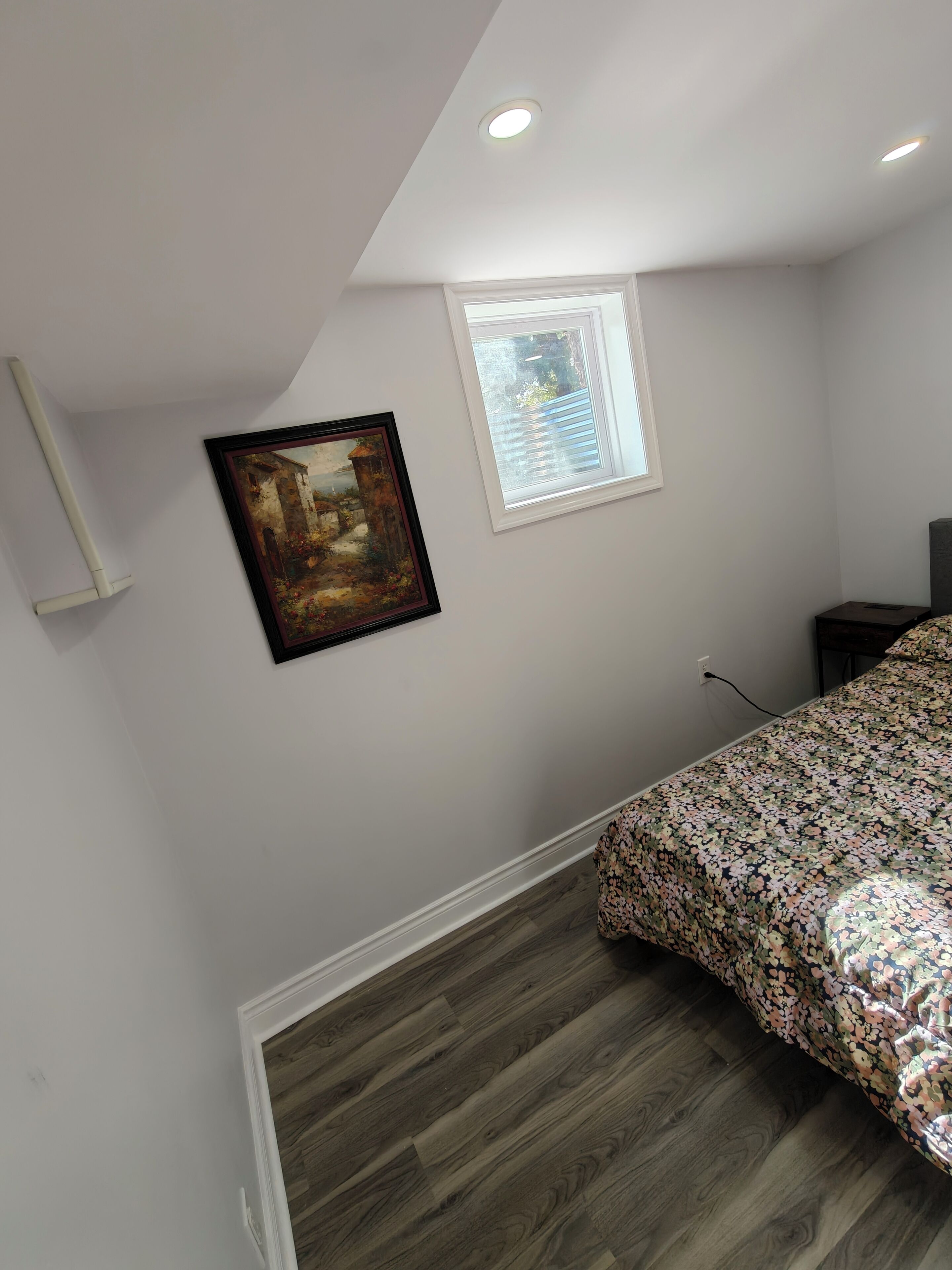 A bedroom with a sloped ceiling, a window with blinds, a framed painting, a bed with a floral bedspread, and a wooden floor.