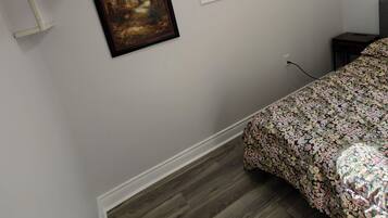 A bedroom with a sloped ceiling, a window with blinds, a framed painting, a bed with a floral bedspread, and a wooden floor.