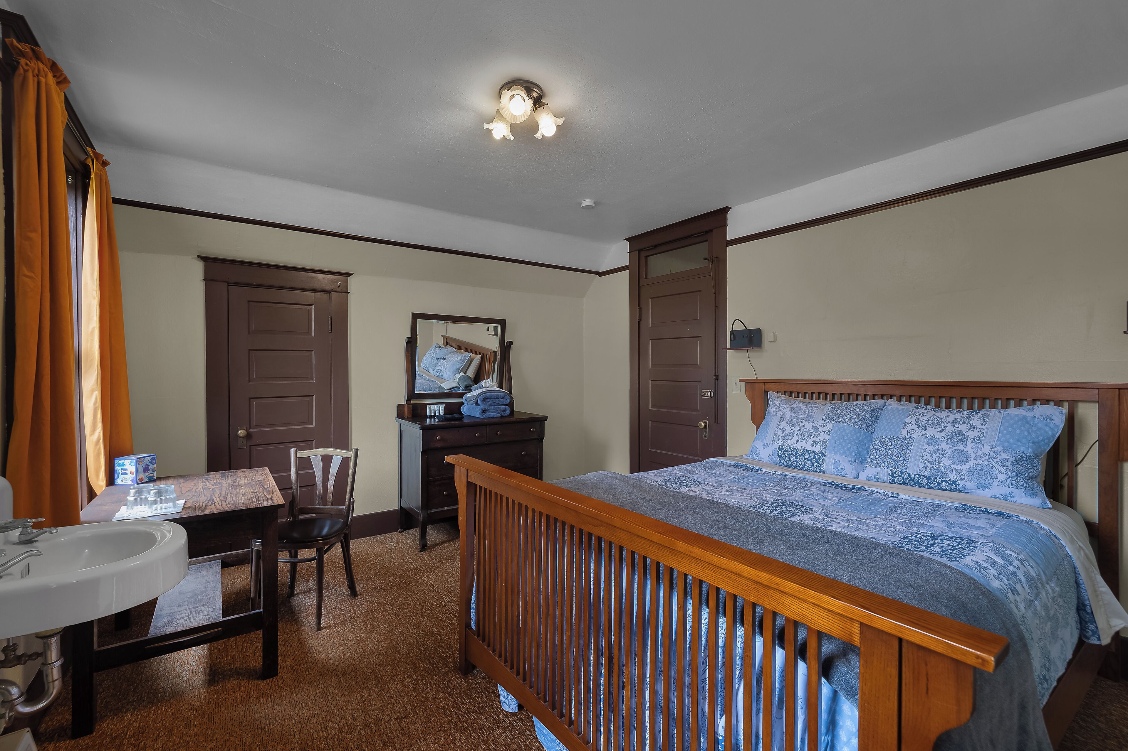 A bedroom with a wooden bed, a dresser, a mirror, a desk with a chair, and a bathroom with a sink and toilet.
