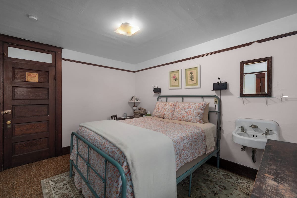 A bedroom with a bed, a wooden door, a bedside table, a nightstand, a wall-mounted mirror, and a wall-mounted sink.
