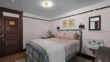A bedroom with a bed, a wooden door, a bedside table, a nightstand, a wall-mounted mirror, and a wall-mounted sink.
