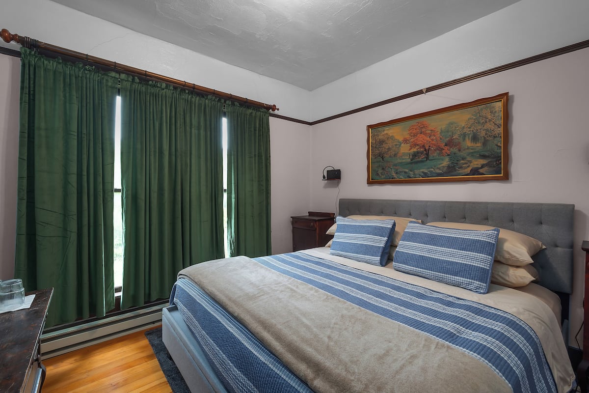 A bedroom with a bed, green curtains, a painting, and a nightstand.