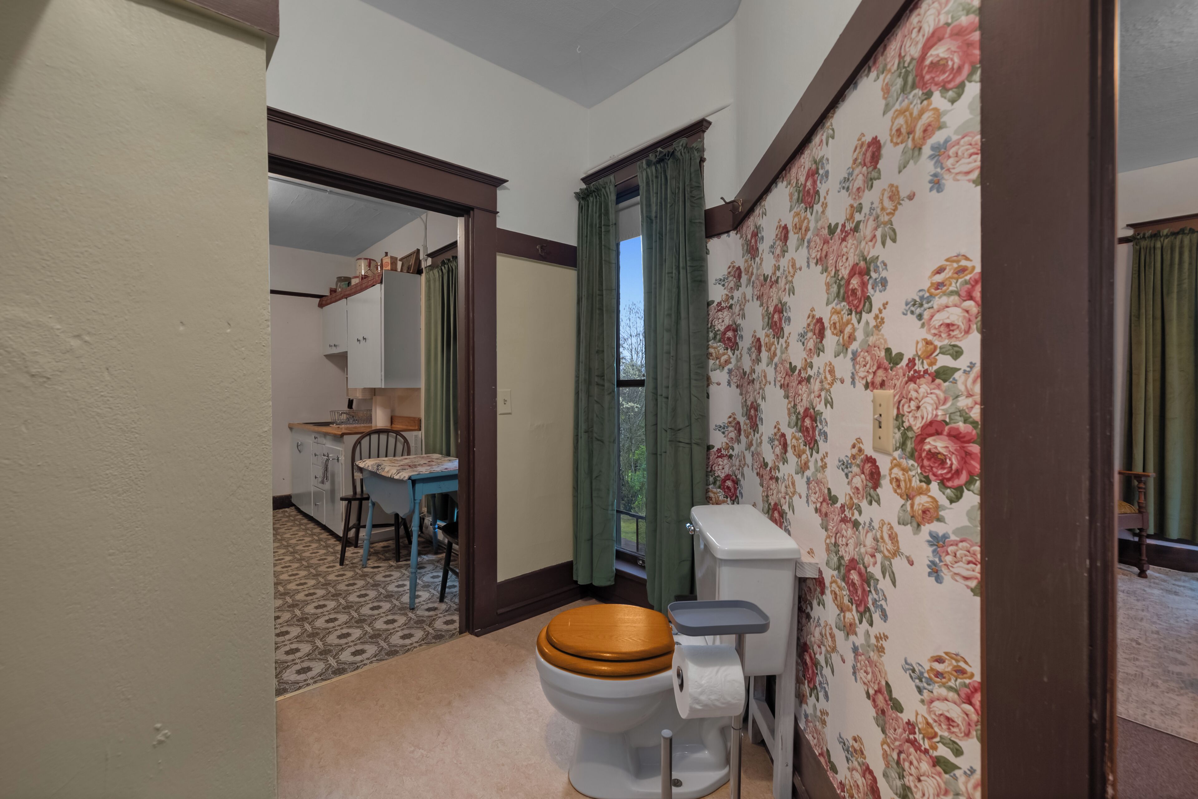 A bathroom with a toilet, wooden seat, and floral wallpaper.
