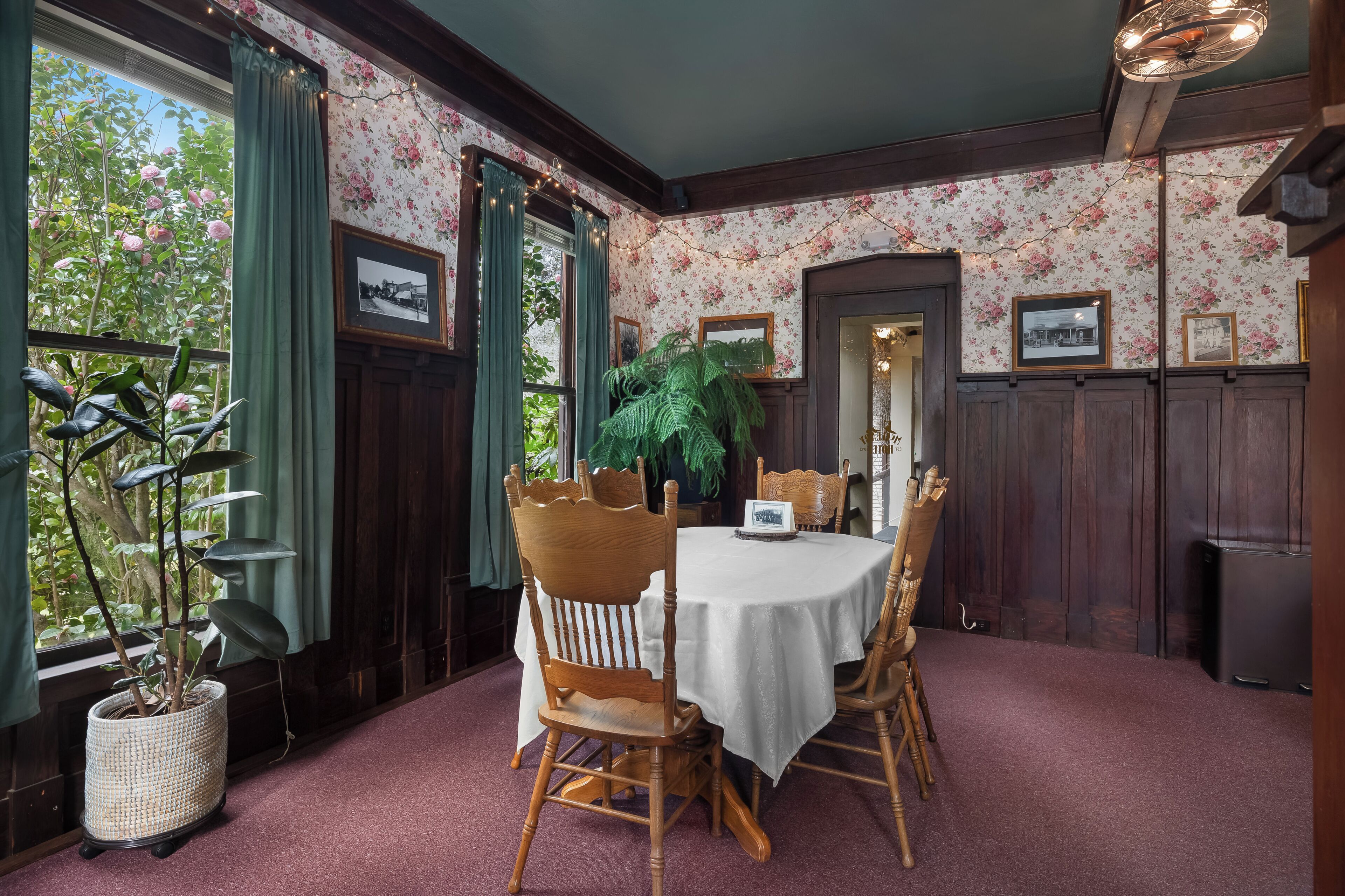 Comedor con una mesa redonda, sillas de madera, una planta en maceta y papel tapiz con motivos florales.