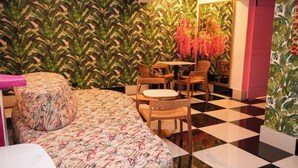 Ein Zimmer mit Schachbrettmuster-Boden, einem Bett mit floralem Überwurf, einem Holztisch mit Stühlen und einer Wand mit tropischem Wandpapier.