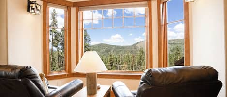 A corner room with two leather chairs, a wooden table with a lamp, and a large window with a view of trees and hills.