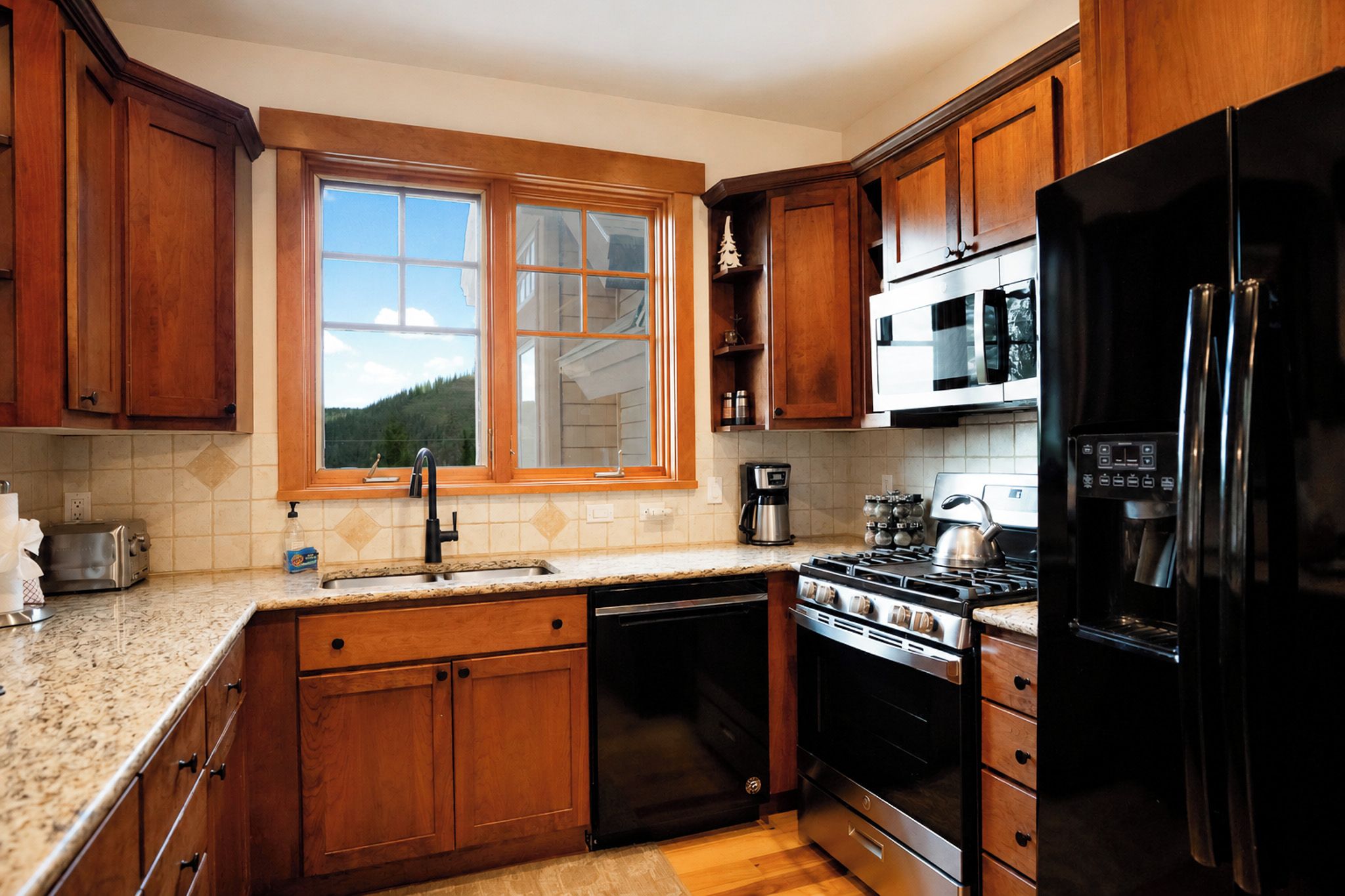 Une cuisine avec des meubles en bois, une cuisinière en acier inoxydable, un réfrigérateur noir et une fenêtre donnant sur une forêt.