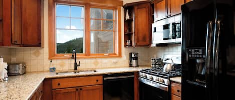 A kitchen with wooden cabinets, a stainless steel stove, a black refrigerator, and a window with a view of a forest.