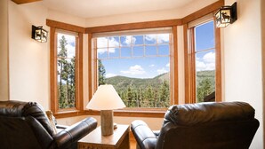 A corner room with two leather chairs, a wooden table, and a lamp. Large windows offer a view of a forest and mountains. - Moonstone Lodge • Hot Tub • Spacious (Breckenridge)