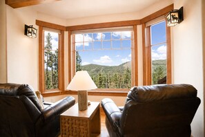 Une pièce d’angle avec deux fauteuils en cuir, une table en bois et une lampe. De grandes fenêtres offrent une vue sur une forêt et des montagnes.