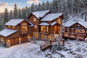 A large log cabin with multiple windows and a stone foundation, surrounded by snow-covered trees.