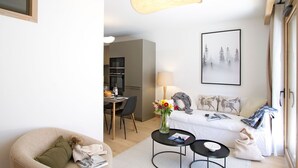 A modern living room with a sofa, a dining table, a kitchen in the background, and a large window. - Ski aux pieds | Piscine & espace bien-être (Alpe d'Huez)