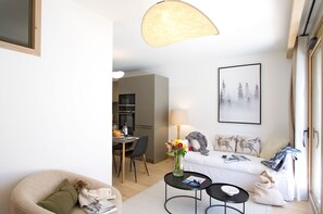 A modern living room with a sofa, a dining table, a kitchen in the background, and a large window. - Ski aux pieds | Piscine & espace bien-être (Alpe d'Huez)