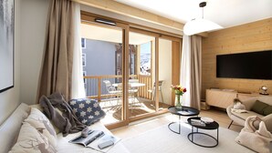 A modern living room with a sofa, coffee table, and TV. There's a balcony with a table and chairs, and a view of a snowy landscape. - Ski aux pieds | Piscine & espace bien-être (Alpe d'Huez)
