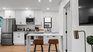 Una cucina moderna con mobili bianchi, forno integrato, microonde e piano cottura. Ci sono due sgabelli e un tavolino.