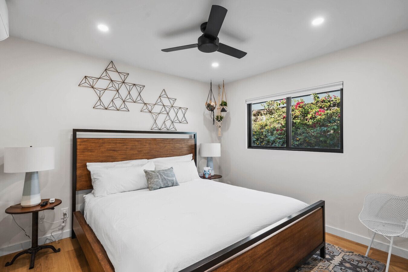 A modern bedroom with a wooden bed, a ceiling fan, a window with a garden view, and decorative wall art.