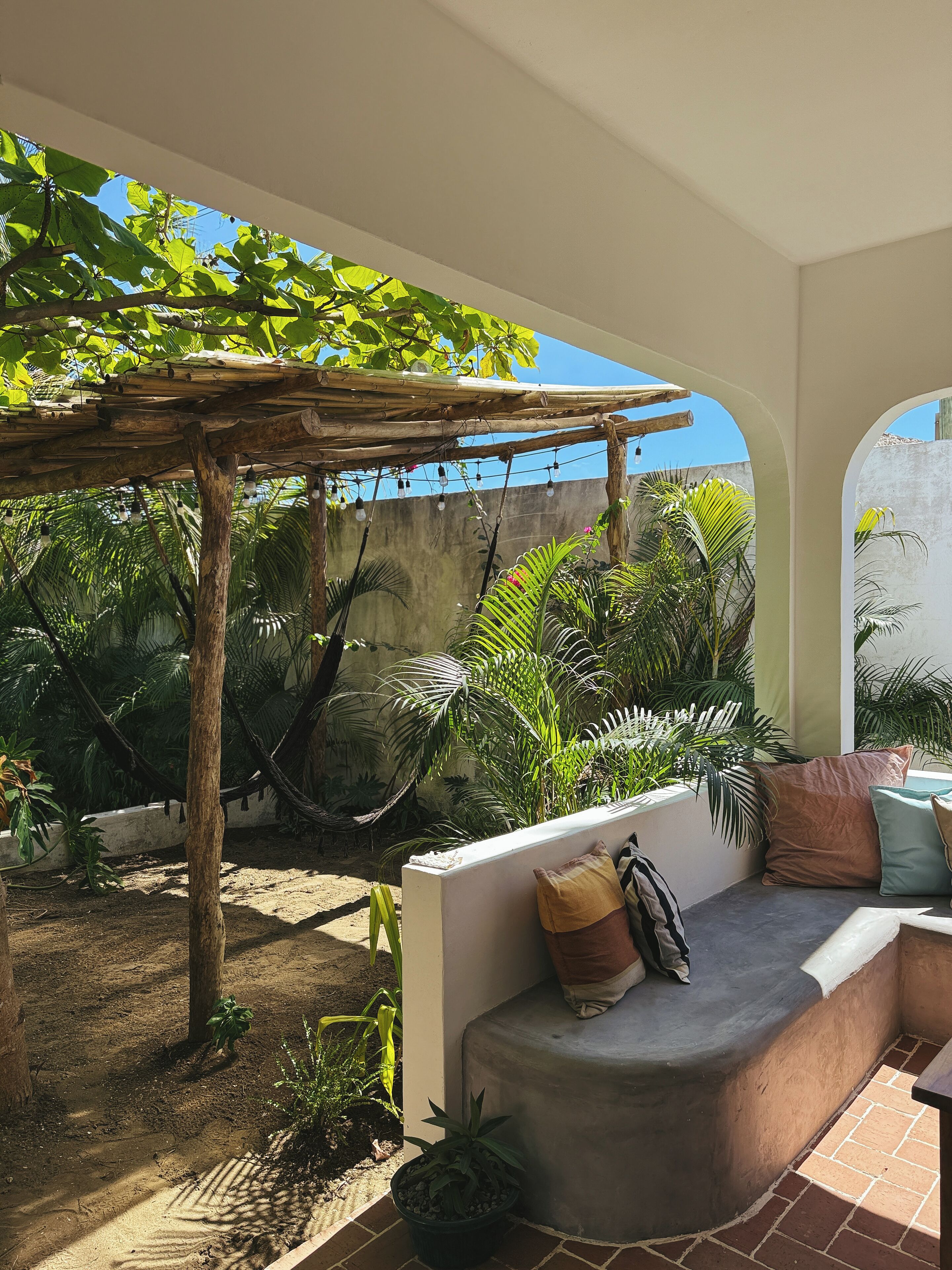 A covered outdoor seating area with a hammock, potted plants, and a concrete bench with cushions.
