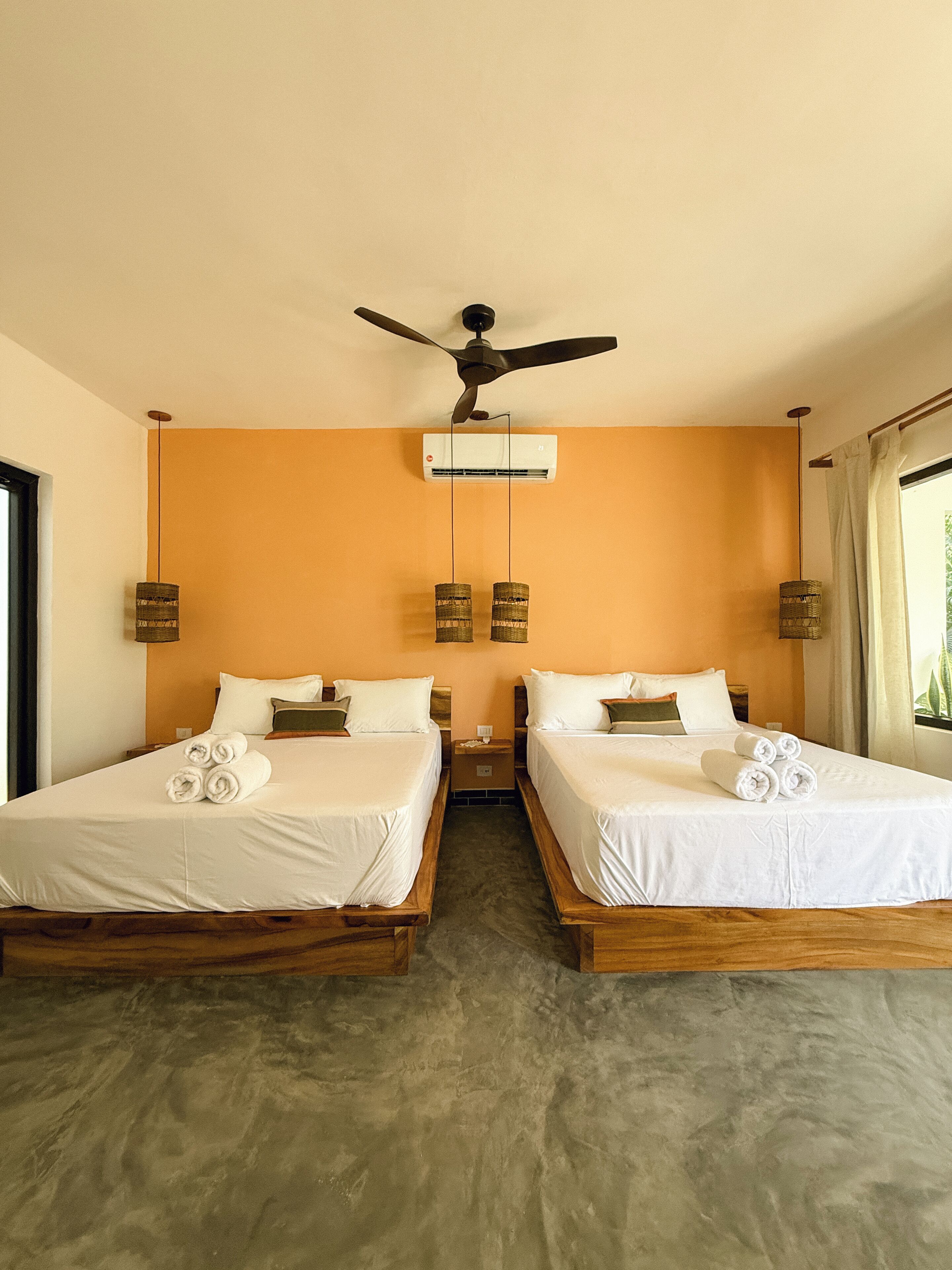 A hotel room with two beds, a ceiling fan, an air conditioning unit, and a window with curtains.