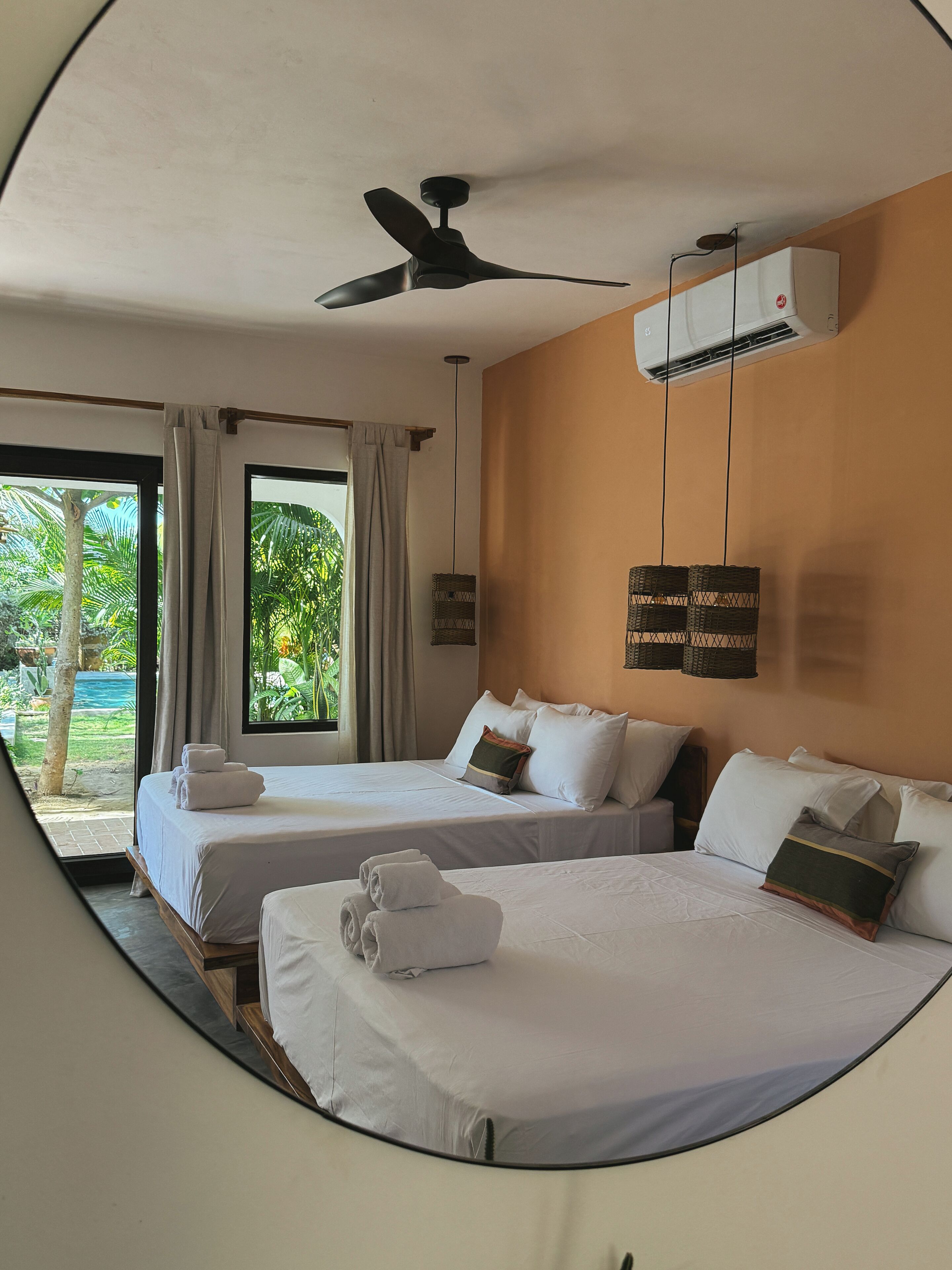 A hotel room with two beds, a ceiling fan, an air conditioning unit, and a view of a tropical garden.