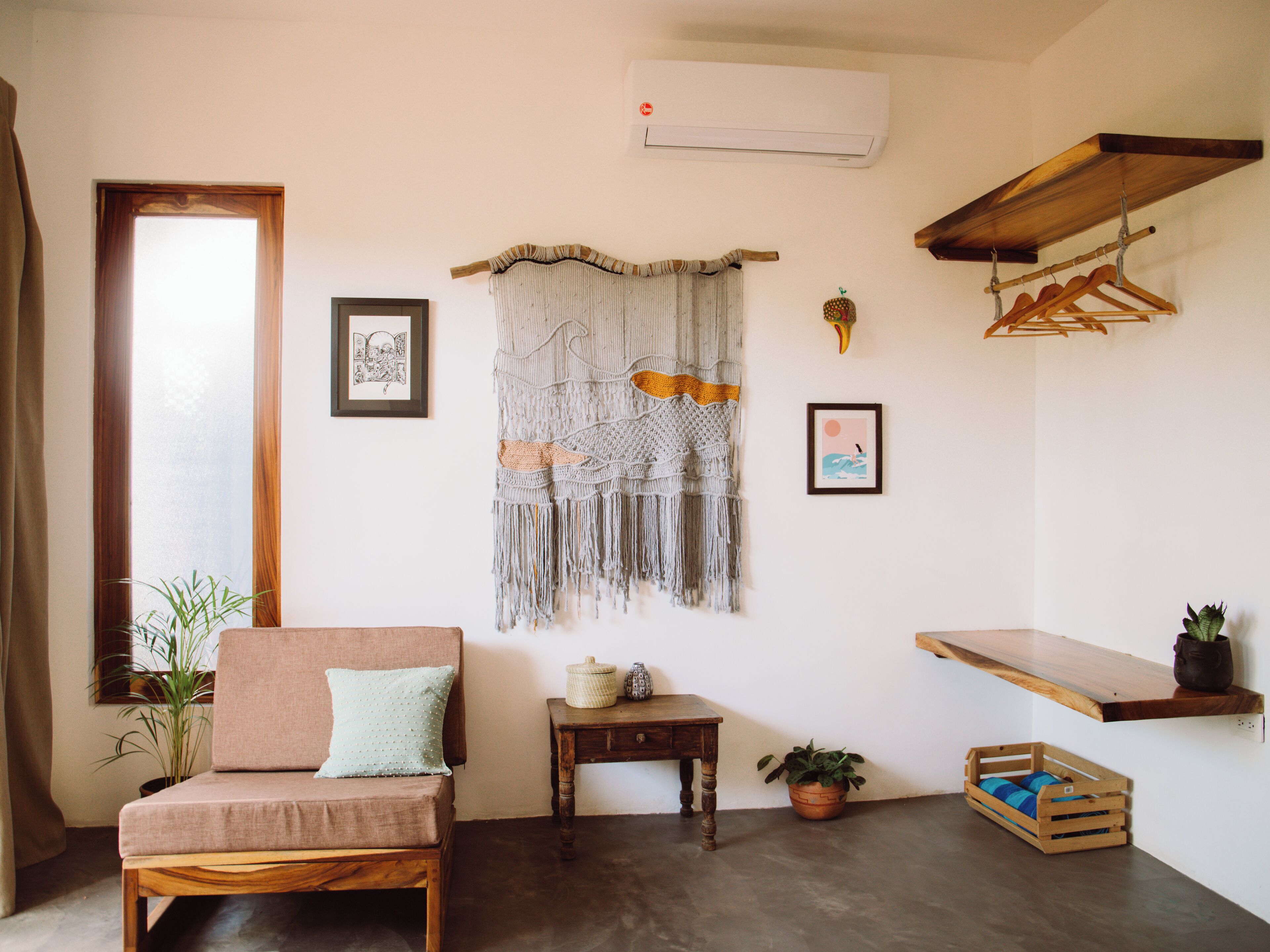 Une pièce chaleureuse avec un canapé en bois, une décoration murale en macramé, une petite table et une étagère en bois ornée d’une plante.