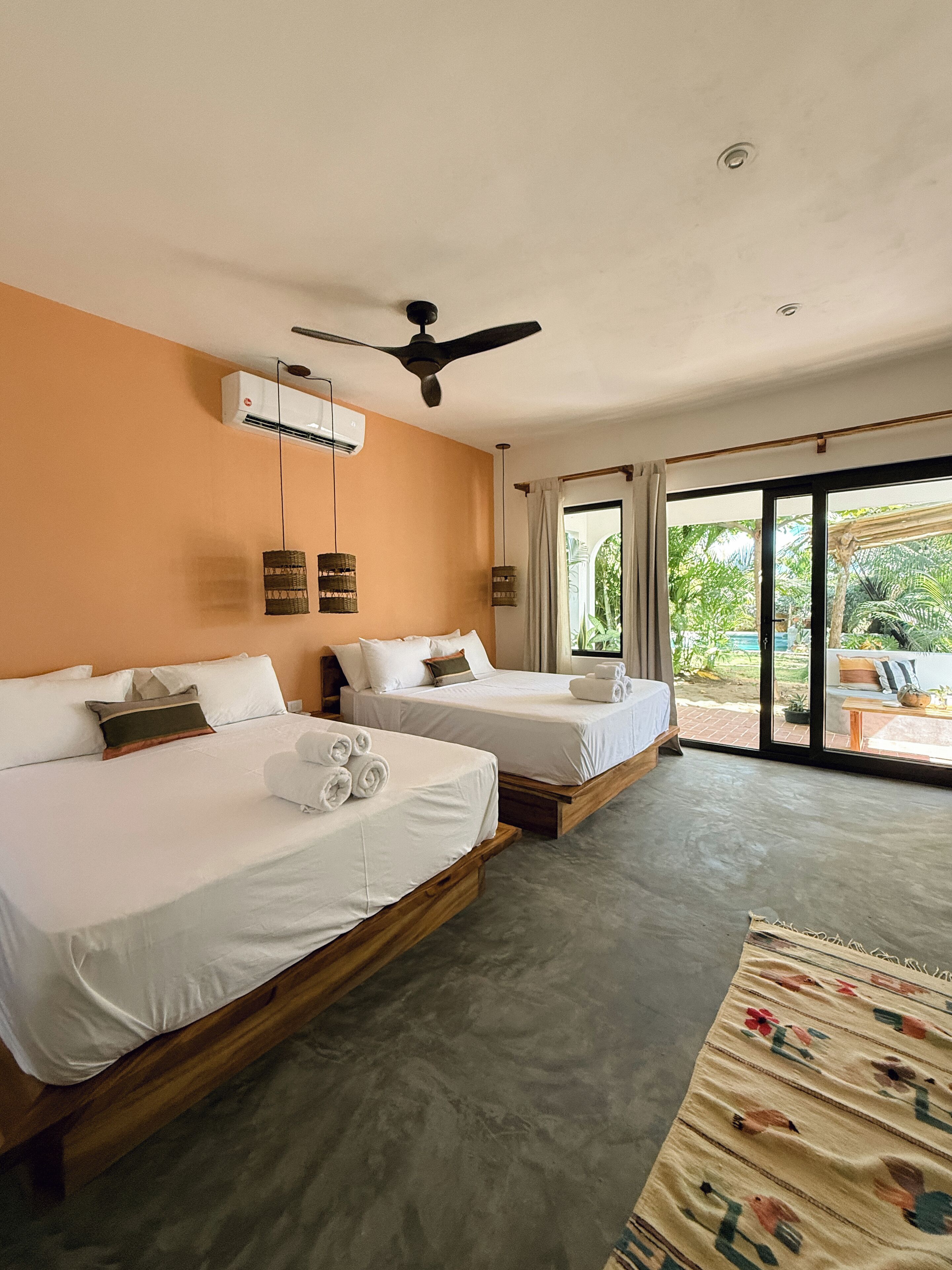 A hotel room with two beds, a ceiling fan, an air conditioning unit, and a view of the outdoors through large sliding doors.