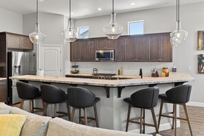 A modern kitchen with a central island, bar stools, stainless steel appliances, and pendant lighting. - The Executive Retreat at Westberry (Madera)