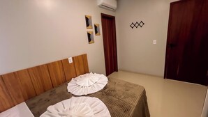 A room with a wooden headboard, two white beds with ruffled bedspreads, a wooden door, and a wall-mounted air conditioner. - Apto com Piscina e 2 su tes a 100m da Praia (Japaratinga)
