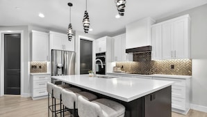 A modern kitchen with white cabinetry, a large island with bar stools, stainless steel appliances, and a herringbone-patterned backsplash. - Oasis at Pecan Valley l Resort Access Included (Hurricane)