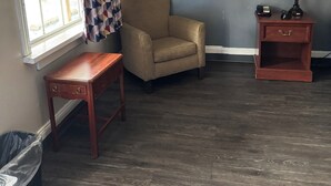 A room with a window, a chair, a small table, a nightstand with a lamp, and a trash bin. - The Executive Inn (Danville)