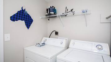 Un espace buanderie comprenant deux machines à laver et deux sèche-linge à chargement frontal, tous deux de couleur blanche, un tissu à carreaux bleus et noirs, et une étagère contenant des produits de nettoyage.